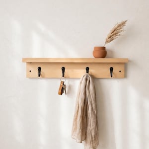 Peut inclure: Une étagère murale en bois clair dotée de quatre crochets en métal noir. Un petit vase couleur terre cuite contenant une tige de plante séchée est posé sur l'étagère. Un trousseau de clés avec un porte-clés en cuir et une écharpe beige sont suspendus.