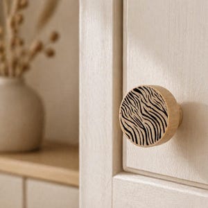 Peut inclure: Un bouton de placard rond en bois avec une finition en bois clair naturel et un motif de rayures zébrées noires imprimé sur la face avant. Le bouton est fixé sur une porte de placard en bois clair, présentant une texture lisse et une forme circulaire.
