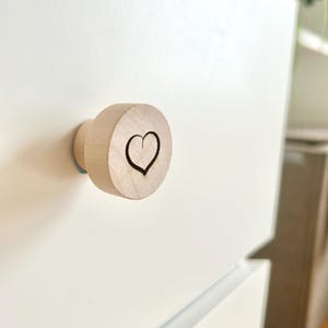 Peut inclure: Un bouton de tiroir en bois avec un motif de cœur gravé à la surface. Le bouton est brun clair et a une finition lisse.