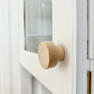 May include: A close-up of a wooden cabinet knob with a round, smooth surface. The knob is attached to a white cabinet door.