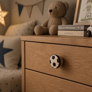 Könnte beinhalten: Eine helle Eichenholzkommode mit einem Fußballknauf. Ein gerahmtes Bild, ein Holzspielzeugauto und Bücher stehen obenauf. Ein Teddybär und ein Sternkissen befinden sich im Hintergrund. Ein sternförmiges Licht hängt an der Wand.