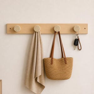 May include: A wooden wall-mounted coat rack with four round, woven knobs. A beige towel, a woven tote bag with brown leather straps, and a set of keys hang from the knobs. The rack is made of light-colored wood.