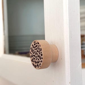 May include: A round, light-colored wooden cabinet knob featuring a flat front face with a dark, laser-engraved leopard print pattern. The knob has a cylindrical base that attaches to a white cabinet door, showcasing a natural wood grain finish.