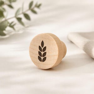 May include: A light-colored wooden knob with a wheat stalk design. The knob is round with a small handle and is made of light-colored wood. The wheat stalk design is dark brown and is centered on the knob.