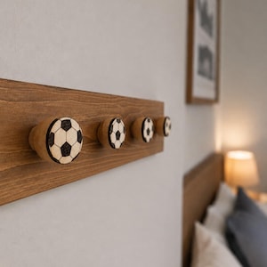 May include: A rectangular wooden wall rack featuring four circular knobs. Each knob is decorated with a black and white football pattern. The rack is made of natural wood with a visible grain, and the knobs are mounted horizontally in a row.
