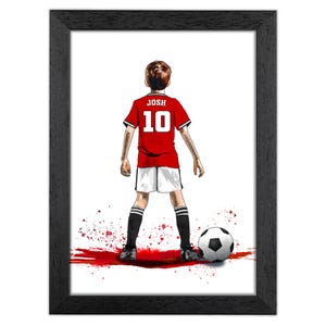 Puede incluir: Impresi&oacute;n art&iacute;stica enmarcada que presenta a un joven jugador de f&uacute;tbol con una camiseta roja con el nombre "JOSH" y el n&uacute;mero "10" en la espalda. El jugador est&aacute; frente a un bal&oacute;n de f&uacute;tbol. La obra es de estilo acuarela con salpicaduras de pintura roja.