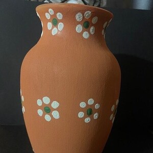 May include: Terracotta-colored vase with a wide body and a flared rim. The vase is adorned with white floral patterns, each with a green center. The handmade vase is suitable for home decor.