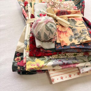 May include: A bundle of assorted floral fabric scraps tied with a beige ribbon. The fabrics feature various floral patterns in shades of red, pink, yellow, and green. A small fabric ball is also included.