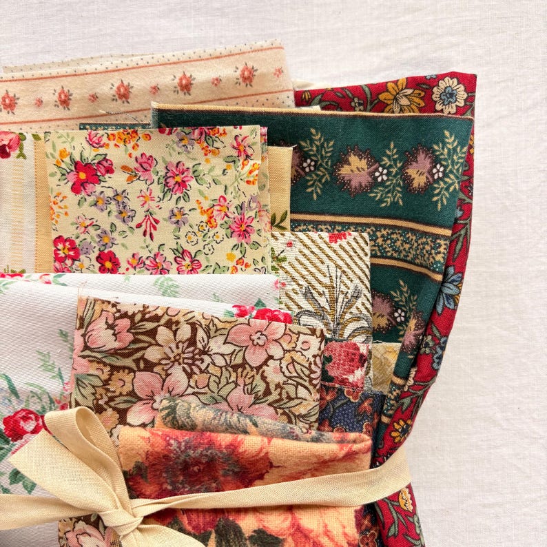May include: A bundle of floral fabric scraps in various patterns and colors, including red, pink, green, and cream. The fabrics are tied together with a beige ribbon, showcasing a variety of floral designs, stripes, and textures. Ideal for quilting or crafting projects.