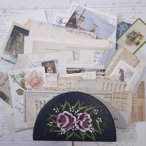 Pode incluir: Uma cole&ccedil;&atilde;o de ef&ecirc;meras vintage, incluindo postais, recibos e cartas manuscritas, dispostas em torno de uma caixa preta em forma de meia-lua. A caixa &eacute; pintada &agrave; m&atilde;o com duas rosas cor-de-rosa e folhas verdes.
