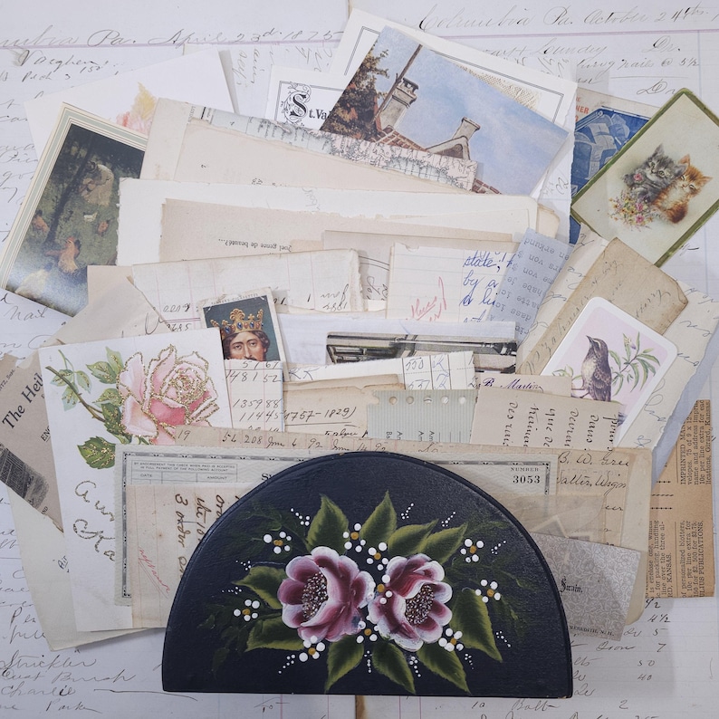 Pode incluir: Uma cole&ccedil;&atilde;o de ef&ecirc;meras vintage, incluindo postais, recibos e cartas manuscritas, dispostas em torno de uma caixa preta em forma de meia-lua. A caixa &eacute; pintada &agrave; m&atilde;o com duas rosas cor-de-rosa e folhas verdes.