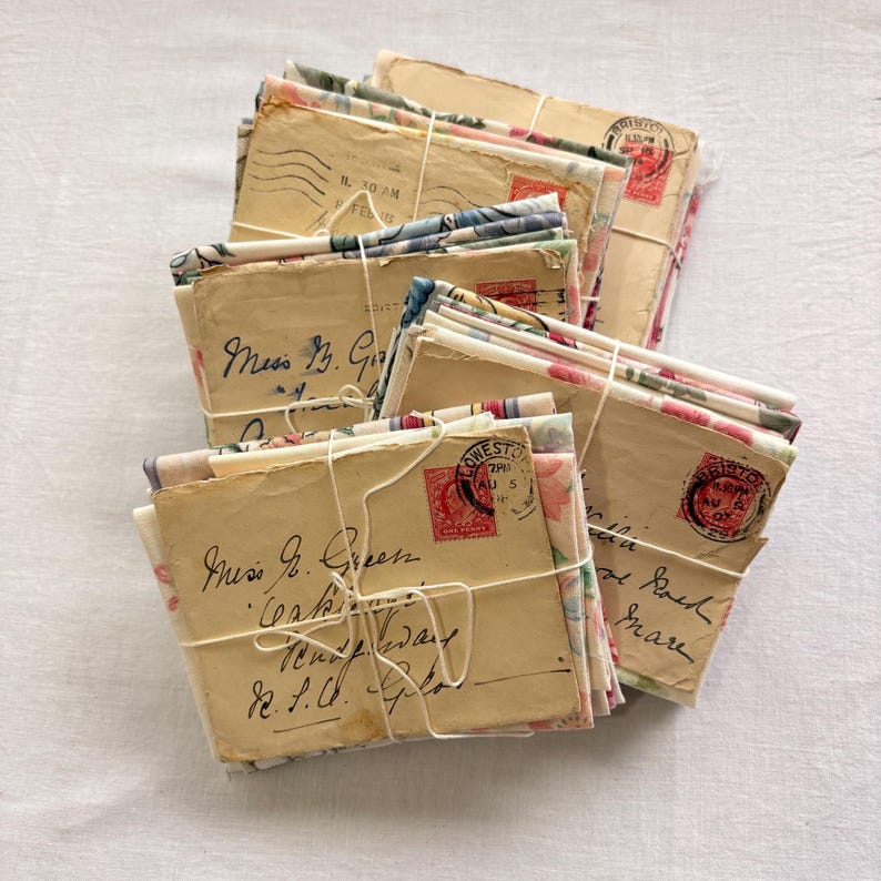 K&ouml;nnte beinhalten: Eine Sammlung alter Briefe, mit Schnur zusammengebunden. Die Umschl&auml;ge sind beige mit handgeschriebenen Adressen und roten Briefmarken. Blumiger Stoff akzentuiert die Briefe und verleiht der antiken Korrespondenz Charme.