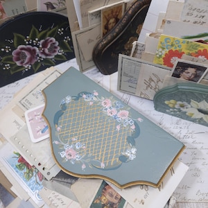 Pode incluir: Uma cole&ccedil;&atilde;o de itens decorativos em estilo vintage. Uma pe&ccedil;a de madeira curva azul clara com um design floral e acabamento dourado est&aacute; em primeiro plano. Outros itens incluem caixas pintadas, cart&otilde;es e pilhas de cartas antigas.
