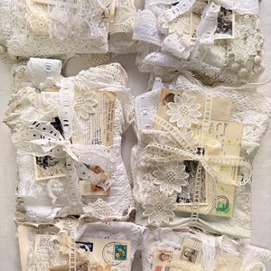 May include: A collection of white lace bundles, each tied with ribbon and containing vintage ephemera. The bundles include postcards, stamps, and decorative lace flowers. The items are arranged on a white surface.
