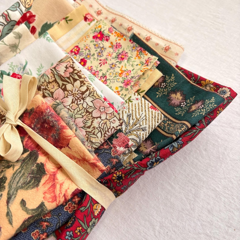 May include: A collection of folded fabric squares with floral patterns in various colors, including red, pink, green, and cream. The fabrics are tied together with a cream-colored ribbon, suggesting a set or bundle of textiles.