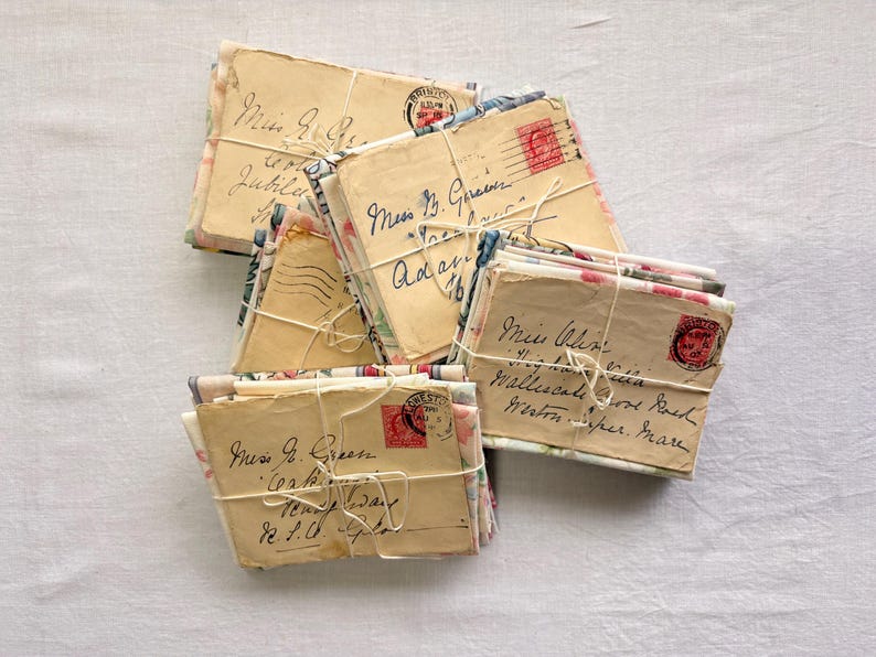 K&ouml;nnte beinhalten: Mehrere B&uuml;ndel antiker Briefe, mit Schnur zusammengebunden. Die Umschl&auml;ge weisen verblasste Handschrift, florale Muster und rote Briefmarken auf. Die Briefe sind auf einer wei&szlig;en Oberfl&auml;che angeordnet, was eine vergangene &Auml;ra andeutet.