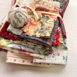May include: A bundle of assorted floral fabric scraps tied with a cream-colored ribbon. The fabrics feature various floral patterns in shades of red, yellow, blue, and green. A ball of fabric with a floral design rests on top of the bundle.