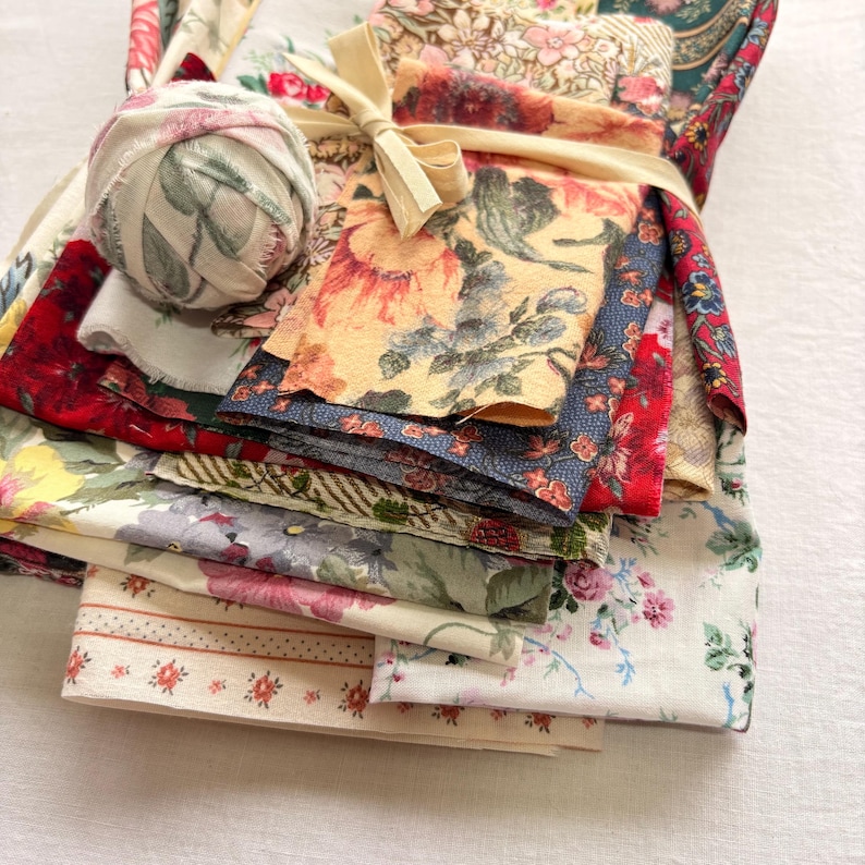 May include: A bundle of assorted floral fabric scraps tied with a cream-colored ribbon. The fabrics feature various floral patterns in shades of red, yellow, blue, and green. A ball of fabric with a floral design rests on top of the bundle.