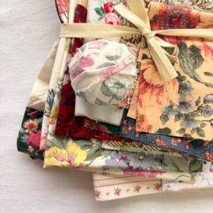 May include: A bundle of floral fabric scraps tied with a cream-colored ribbon. The fabrics feature various floral patterns in shades of red, yellow, pink, and blue. A small fabric ball is also included in the bundle. The fabrics are stacked on top of each other.