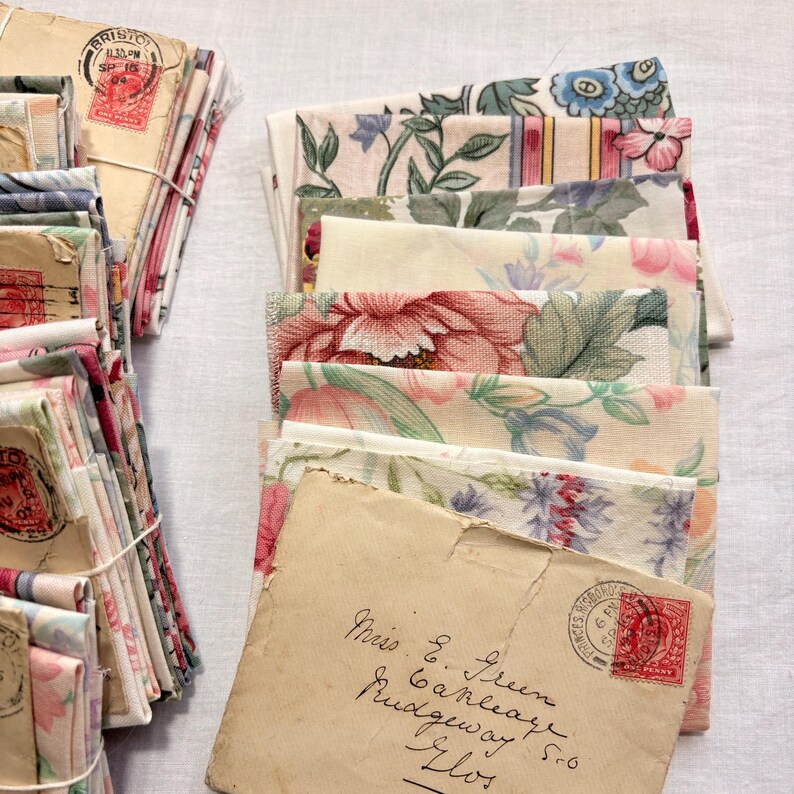 K&ouml;nnte beinhalten: Stapel von Vintage-Stoffresten und Umschl&auml;gen mit Briefmarken. Der Stoff weist florale Muster in Rosa, Blau und Gr&uuml;n auf. Die Umschl&auml;ge haben handschriftliche Adressen und Poststempel.
