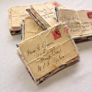 K&ouml;nnte beinhalten: Mehrere Stapel alter Briefe, die mit Bindfaden zusammengebunden sind. Die Umschl&auml;ge sind gealtert und weisen handschriftliche Adressen und Poststempel auf. Einige Umschl&auml;ge haben florale Muster. Die Briefe sind mit wei&szlig;em Bindfaden gebunden.