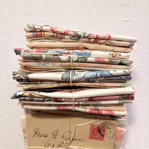 K&ouml;nnte beinhalten: Ein Stapel gefalteter Stoffe, mit Schnur gebunden. Der Stoff zeigt florale Muster in Rosa, Blau und Gr&uuml;n auf cremefarbenem Hintergrund. Darunter befinden sich Vintage-Umschl&auml;ge mit Handschrift und Briefmarken.