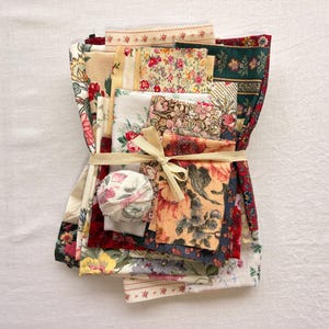 May include: A bundle of assorted fabric scraps tied with a cream-colored ribbon. The fabrics feature floral and patterned designs in various colors, including red, yellow, green, and cream. A small, round fabric item is also included in the bundle.