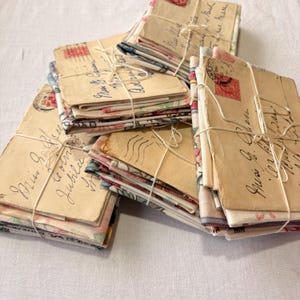 K&ouml;nnte beinhalten: Mehrere Stapel Vintage-Umschl&auml;ge, mit Bindfaden gebunden. Die Umschl&auml;ge sind beige mit handschriftlichen Schriftz&uuml;gen und Briefmarken. Blumig gemusterter Stoff ist an den R&auml;ndern zu sehen. Ein nostalgisches und romantisches Thema.