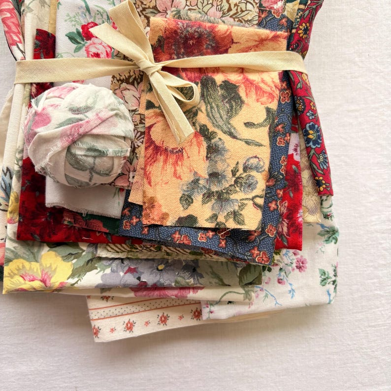 May include: A bundle of assorted floral fabric scraps tied with a cream-colored ribbon. The fabrics feature various floral patterns in red, yellow, blue, and white. A small fabric ball is also included in the bundle.