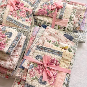 May include: A collection of floral-patterned quilts and smaller quilted pieces, tied with pink ribbons. The quilts display diverse floral patterns in pink, blue, and green on a cream backdrop, with some having blue borders.