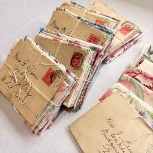 K&ouml;nnte beinhalten: Gestapelte Vintage-Umschl&auml;ge, mit floralen Stoffeins&auml;tzen, mit Schnur gebunden. Die Umschl&auml;ge sind gealtert und tragen handgeschriebene Adressen und rote Briefmarken. Das Bild hat ein nostalgisches, antikes Thema.