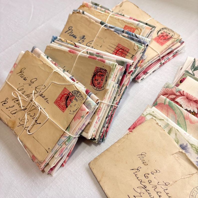 K&ouml;nnte beinhalten: Gestapelte Vintage-Umschl&auml;ge, mit floralen Stoffeins&auml;tzen, mit Schnur gebunden. Die Umschl&auml;ge sind gealtert und tragen handgeschriebene Adressen und rote Briefmarken. Das Bild hat ein nostalgisches, antikes Thema.