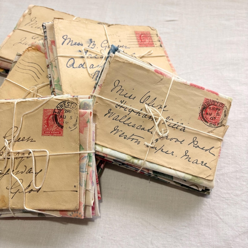 K&ouml;nnte beinhalten: Mehrere B&uuml;ndel alter Briefe, die mit Bindfaden zusammengebunden sind. Die Umschl&auml;ge sind beige und tragen handschriftliche Adressen und rote Briefmarken. Einige Umschl&auml;ge haben Stoff mit Blumenmuster. Der Text enth&auml;lt Namen und Adressen.
