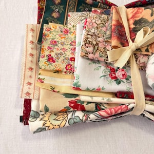 May include: A bundle of assorted fabric squares tied with a cream-colored ribbon. The fabrics feature floral patterns in various colors, including red, pink, yellow, and green. Some fabrics have stripes or other decorative elements. The bundle is on a white surface.