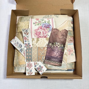 Puede incluir: Una caja de cartón abierta llena de muestras de papel pintado vintage. Las muestras presentan diseños florales en rosa, azul y crema, junto con tiras decorativas. La caja revela la variedad de diseños.