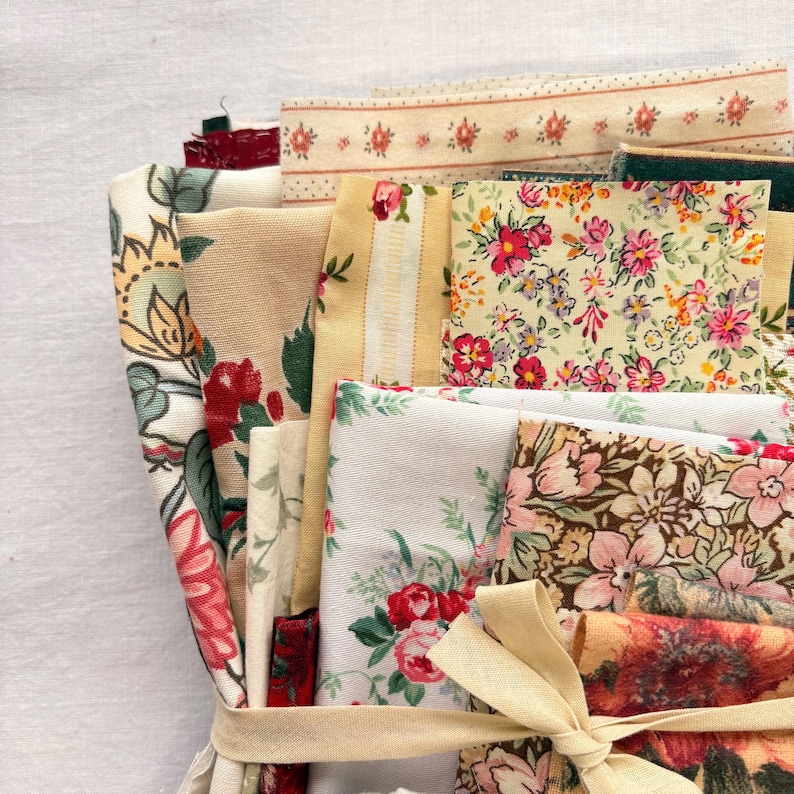 May include: A bundle of assorted fabric swatches in various floral patterns and colors, including red, pink, yellow, and green. The fabrics are tied together with a beige ribbon. The bundle includes a variety of textures and designs, perfect for crafting or sewing projects.