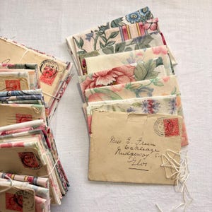 K&ouml;nnte beinhalten: Stapel alter Umschl&auml;ge und Stoffreste mit Blumenmuster. Die Umschl&auml;ge sind beige mit roten Briefmarken und handschriftlichen Adressen. Die Stoffst&uuml;cke zeigen florale Muster in Rosa, Blau und Gr&uuml;n. Mit Schnur gebunden.