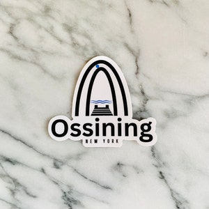 May include: A white sticker with a black outline of an archway with a blue river and a black bridge inside. The text "Ossining NEW YORK" is below the archway.