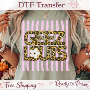 May include: A sage green t-shirt featuring the text "GEEZ LOUIS" in a leopard print design, set against a pink striped background. A white flower graphic is incorporated into the design. The words "DTF Transfer" are at the top.