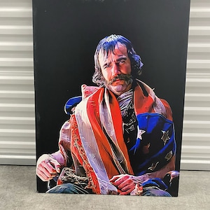 Può includere: Un uomo con la barba e i capelli lunghi è avvolto in una bandiera americana. È seduto su una sedia e guarda la telecamera. Lo sfondo è nero.