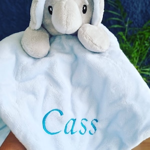 May include: A light blue baby blanket with a gray elephant plush toy attached. The blanket has the name "Cass" embroidered on it in turquoise thread.