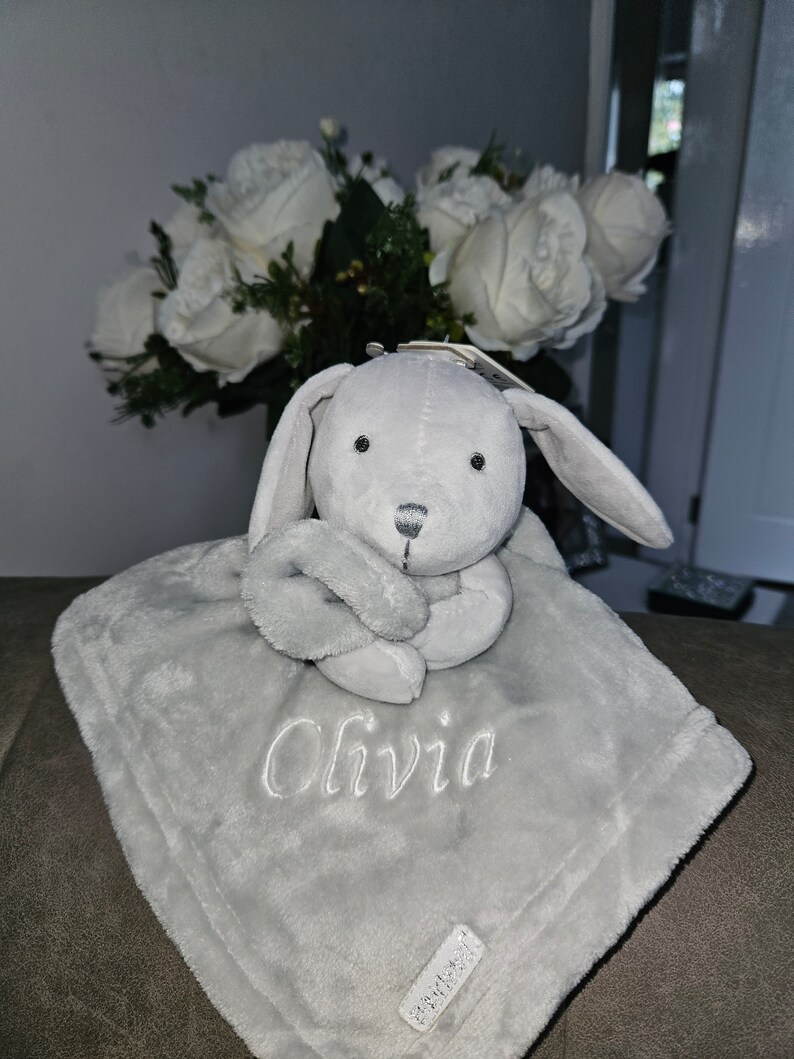 May include: A gray plush bunny with a gray blanket. The blanket has the name "Olivia" embroidered on it.