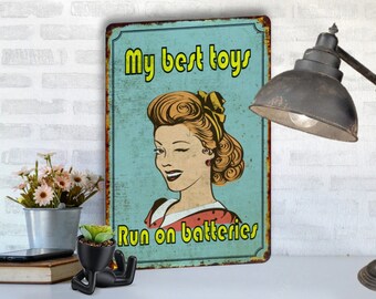 May include: A vintage-style metal sign with a blue background and a cartoon image of a woman winking. The text on the sign reads "My best toys run on batteries".