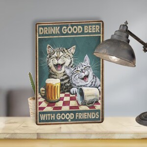 May include: A vintage-style metal sign with a green background featuring two cats laughing and holding drinks. The text on the sign reads "Drink good beer with good friends".