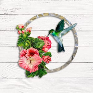 May include: A metal circle with a silver finish, featuring a hummingbird with green and blue feathers, hovering near pink flowers. The background is a white woodgrain surface.