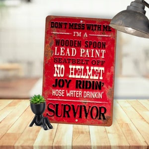 May include: Red metal sign with a distressed, rusty look. The sign reads "Don't mess with me, I'm a wooden spoon, lead paint, seatbelt off, no helmet, joy ridin', hose water drinkin' survivor".