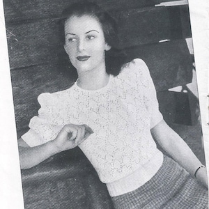 Op de afbeelding: Zwart-witfoto van een vrouw die een witte gehaakte trui draagt met een sterpatroon. De trui heeft pofmouwen en een ronde hals. De vrouw leunt tegen een houten oppervlak. De tekst "Tell Tale Knit" is zichtbaar in de rechterbovenhoek van de afbeelding. De tekst "Design No. 2430" is zichtbaar in de rechterbenedenhoek van de afbeelding.