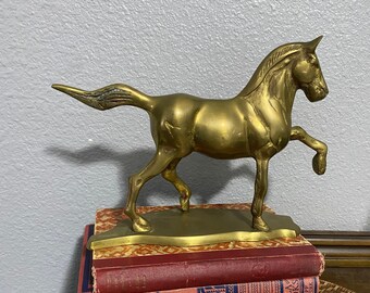 May include: A gold-colored brass horse figurine standing on a stack of three vintage books. The books have red and blue covers with gold lettering. The horse is in a running pose with its head turned to the side.