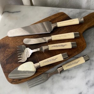 May include: A vintage serving set with five pieces, including a cake server, knife, fork, and two other utensils. The handles are cream-colored with a silver floral design, and the blades are stainless steel. The set is arranged on a wooden cutting board.