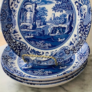 May include: A stack of three blue and white ceramic plates with a detailed landscape design. The plates are stacked on a gold-colored metal plate holder. The plates have a decorative floral border.